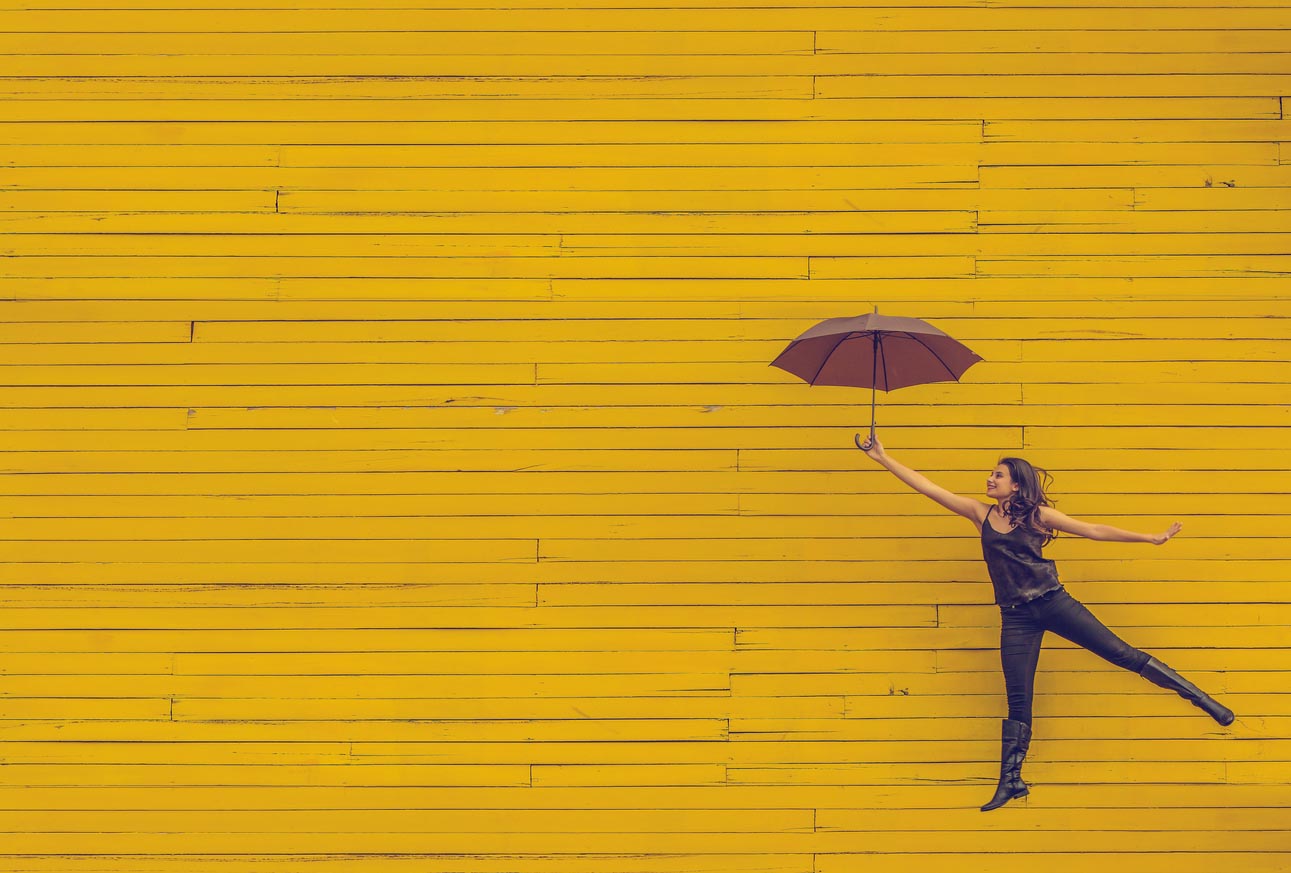 Woman with parasol jumping against yellow wall